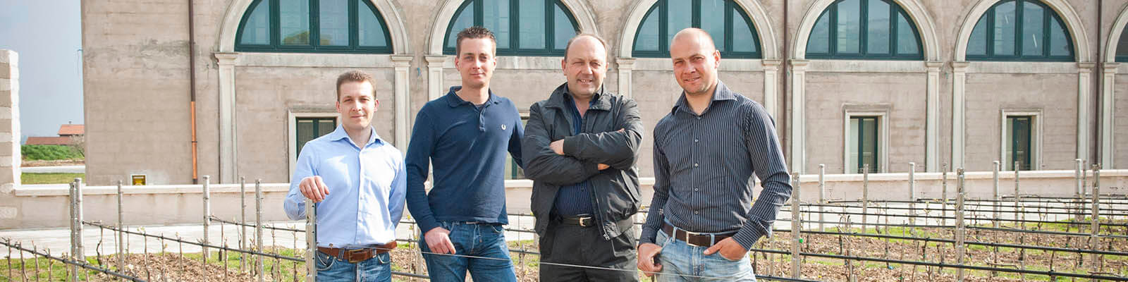 Four men are standing outdoors in front of a building with arched windows. They are on a patio with young plants in rows. They are casually dressed and smiling at the camera.