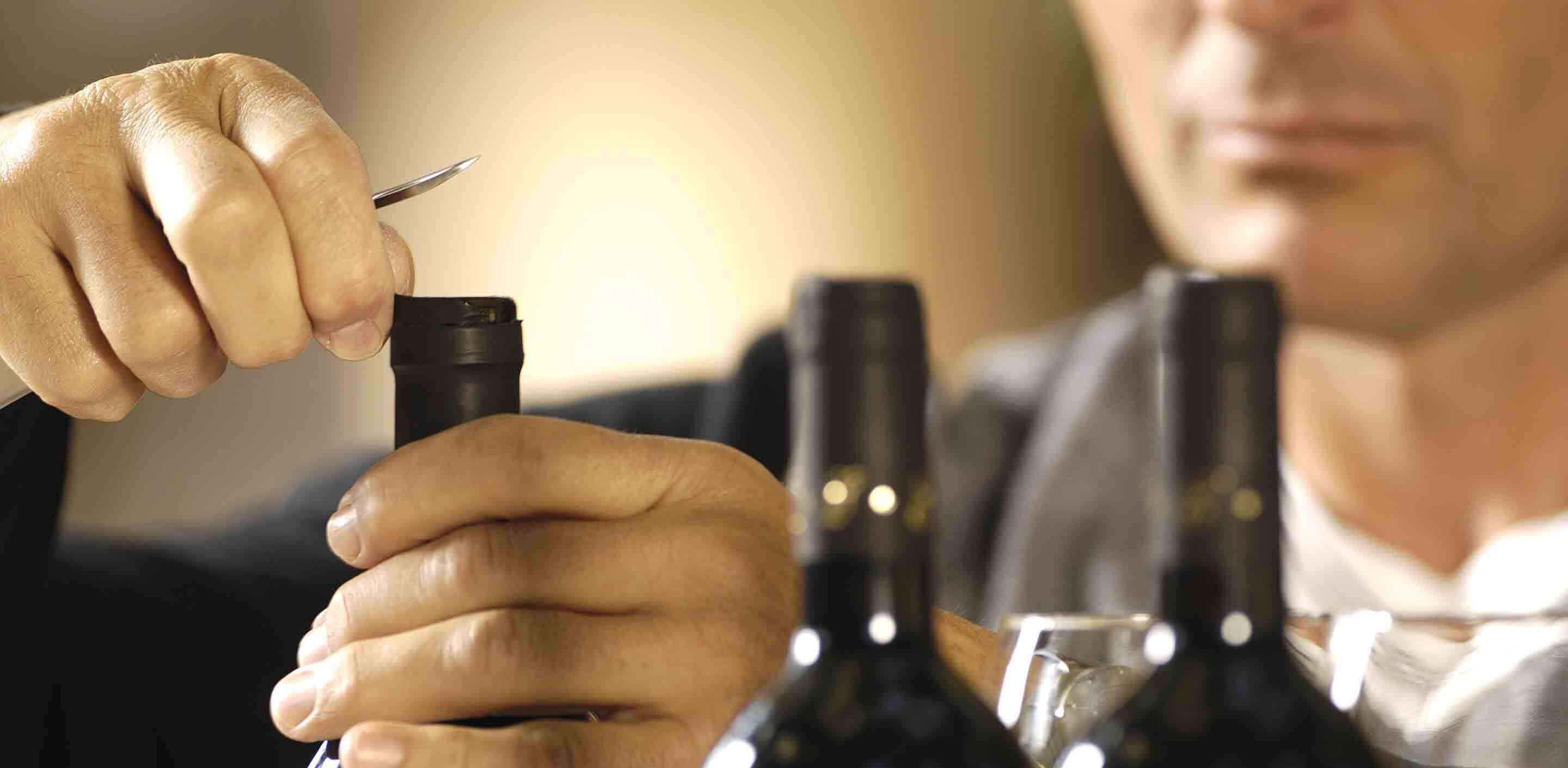 A person using a corkscrew to open a wine bottle, with a blurred background. Two other wine bottles are partially visible in the foreground. The focus is on the hands and the bottle being opened.