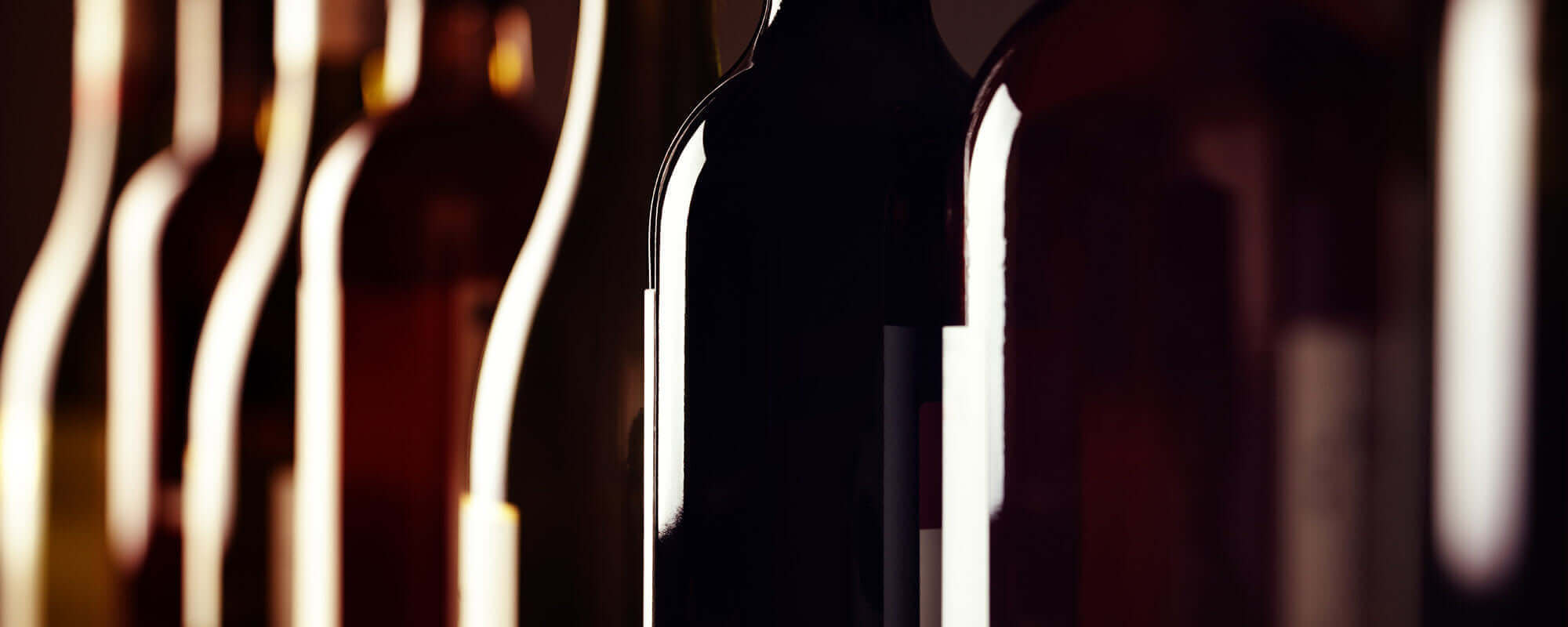 Close-up of several wine bottles in dim lighting, highlighting their silhouettes. The bottles vary in color and create an elegant, moody atmosphere with reflections along their curves.