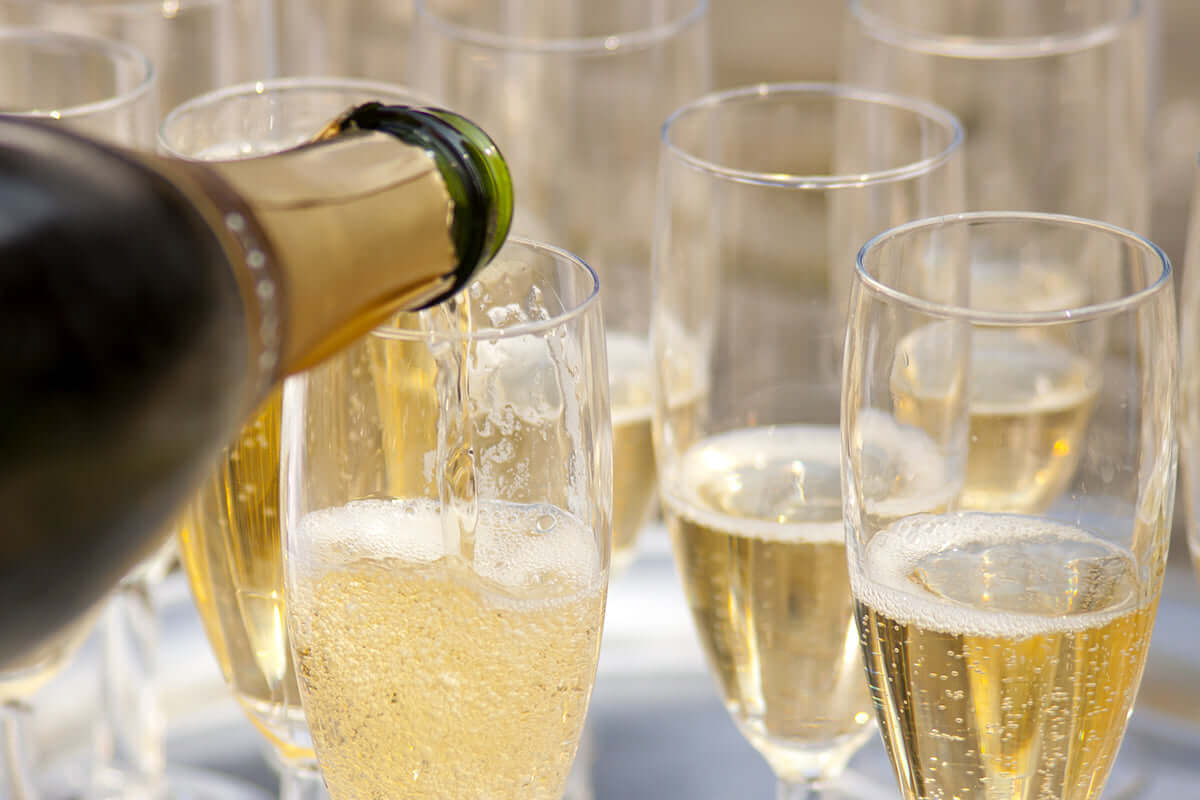 Close-up of a bottle pouring champagne into a glass, surrounded by several other filled flutes. The golden liquid is bubbling and sparkling, suggesting a celebratory occasion. The background is softly blurred.