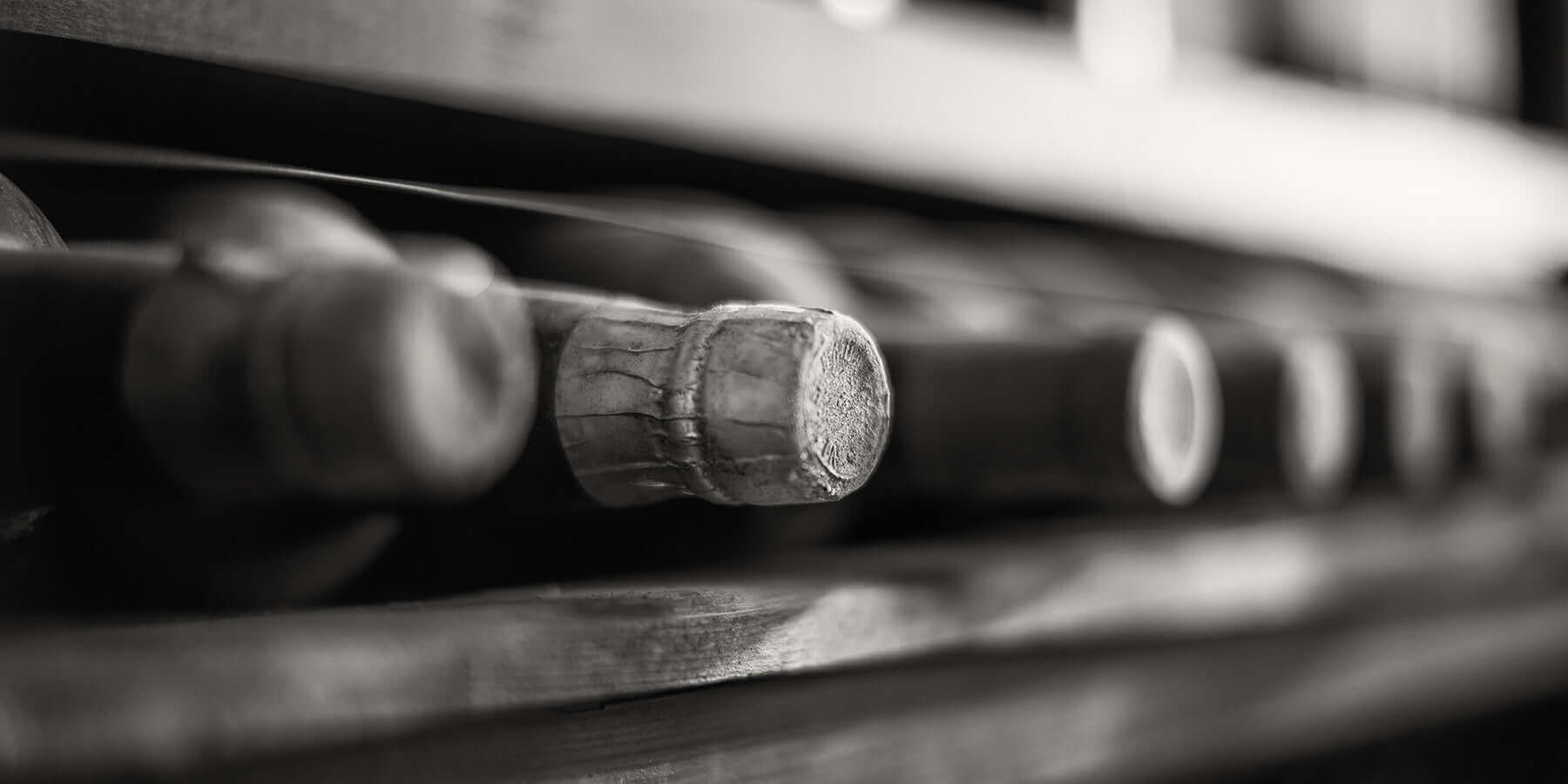 Close-up, black and white image of several wine bottles stored horizontally on a wooden rack. The focus is on the corks, creating a rustic and vintage atmosphere.