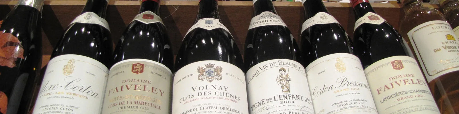 A row of bottles of red wine displayed on a shelf. The labels include names like Domaine Faiveley, Volnay Clos des Chênes, and Pommard Premier Cru. The bottles vary in vintage and origin.