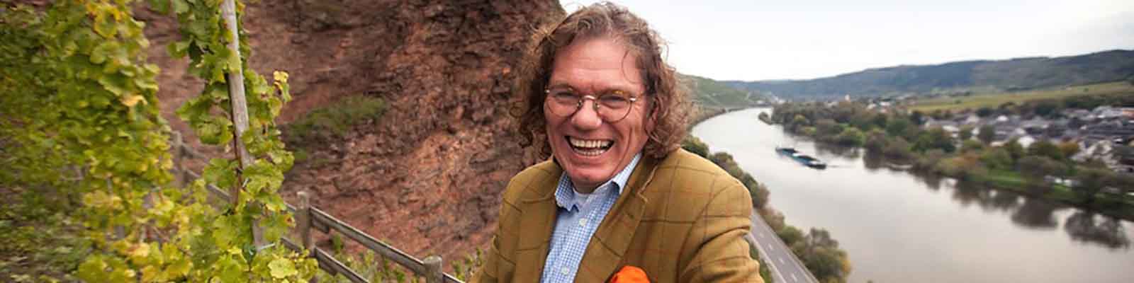 A man with long hair and glasses smiles broadly while standing outdoors. Behind him, a river winds through a lush, hilly landscape with trees and a rocky cliff. He is wearing a brown jacket and blue shirt.