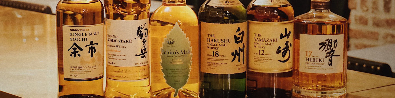 A row of assorted whiskey bottles with various labels, including Yoichi, Hakushu, Yamazaki, and Ichiros Malt, displayed on a wooden surface.