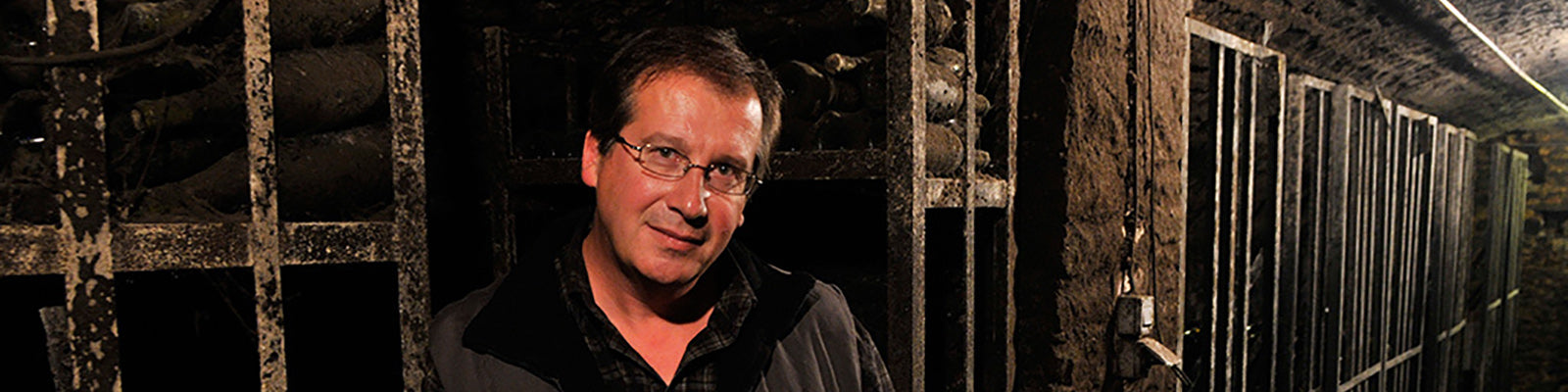 A person with glasses stands in a dimly lit cellar lined with metal racks and stacks of bottles. The atmosphere is rustic and the lighting is warm.
