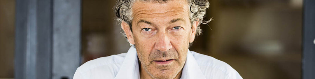 An older man with grey hair is looking directly at the camera. He is wearing a white shirt and has a neutral expression. The background is blurred, possibly depicting a room or outdoor setting.