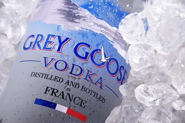 A bottle of Grey Goose vodka surrounded by ice cubes. The label reads Distilled and Bottled in France with a small French flag. The design features a flying goose.