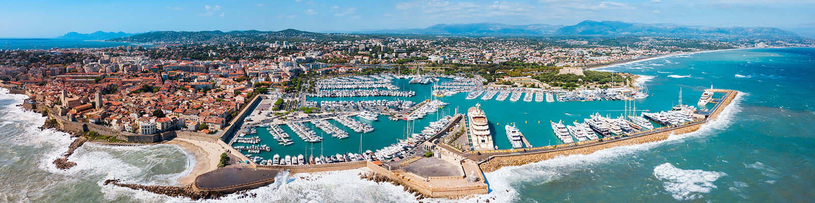 A panoramic view of a coastal town with a large marina filled with yachts. The town features a mix of historic and modern buildings, bordered by a stone breakwater. Blue sea and waves are visible, with a backdrop of lush green hills under a clear sky.