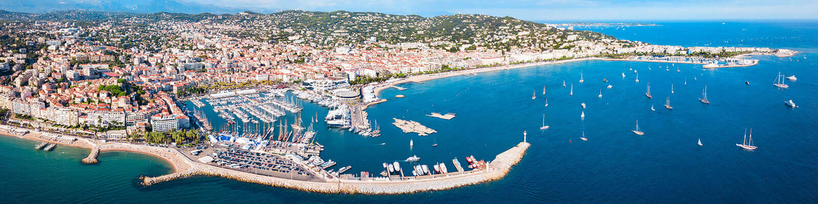 Aerial view of a coastal city featuring a marina filled with yachts and sailboats. The cityscape is densely packed with buildings, surrounded by green hills. The azure sea stretches to the horizon under a clear blue sky.