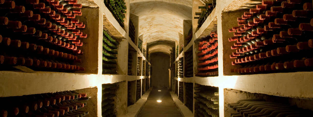 A dimly lit wine cellar with rows of shelves stacked with wine bottles on either side of a narrow passage. The bottles are laid horizontally, and the stone walls create an old-world ambiance.