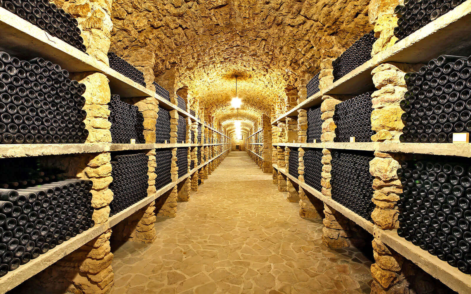 A long, stone wine cellar with arched ceilings, lined with shelves packed with numerous bottles. The dim lighting casts a warm glow, highlighting the textured walls and floor, creating an inviting, rustic ambiance.
