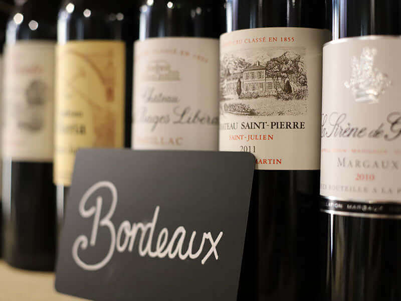 Close-up of several Bordeaux wine bottles lined up on a shelf. A black card with Bordeaux written in white cursive is in the foreground. The bottle labels display various winery names and vintages.