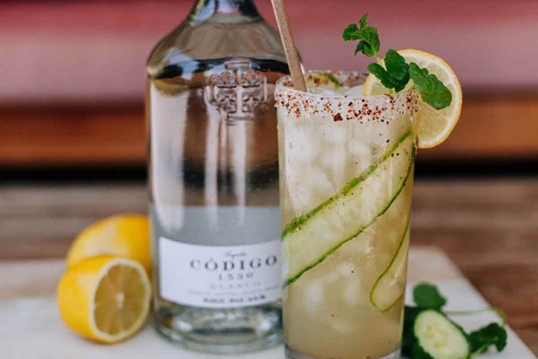 A refreshing cocktail garnished with cucumber slices, lemon, and mint is served in a tall glass with a salted rim. A bottle of Código 1530 tequila is beside the drink, along with two lemons and mint leaves on a marble surface.