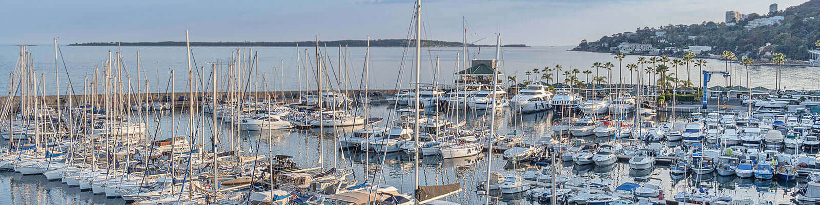 A panoramic view of a marina filled with numerous sailboats and yachts docked alongside piers. In the background, a calm sea stretches towards a distant shoreline and a row of houses on a hillside, under a partly cloudy sky.