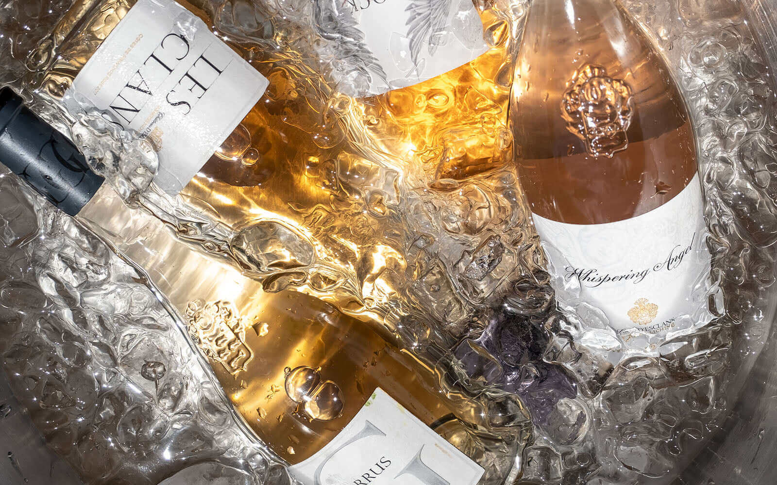 Bottles of chilled white and rosé wine submerged in ice, showcasing labels including Les Clans and Whispering Angel. The bottles are partially obscured by the ice, creating a refreshing and inviting scene.