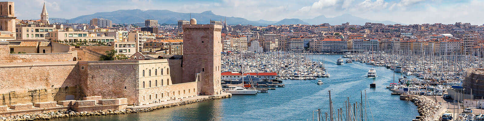 A scenic view of a bustling harbor filled with numerous boats, bordered by historic stone buildings on the left and a vibrant cityscape with mountains in the background under a partly cloudy sky.