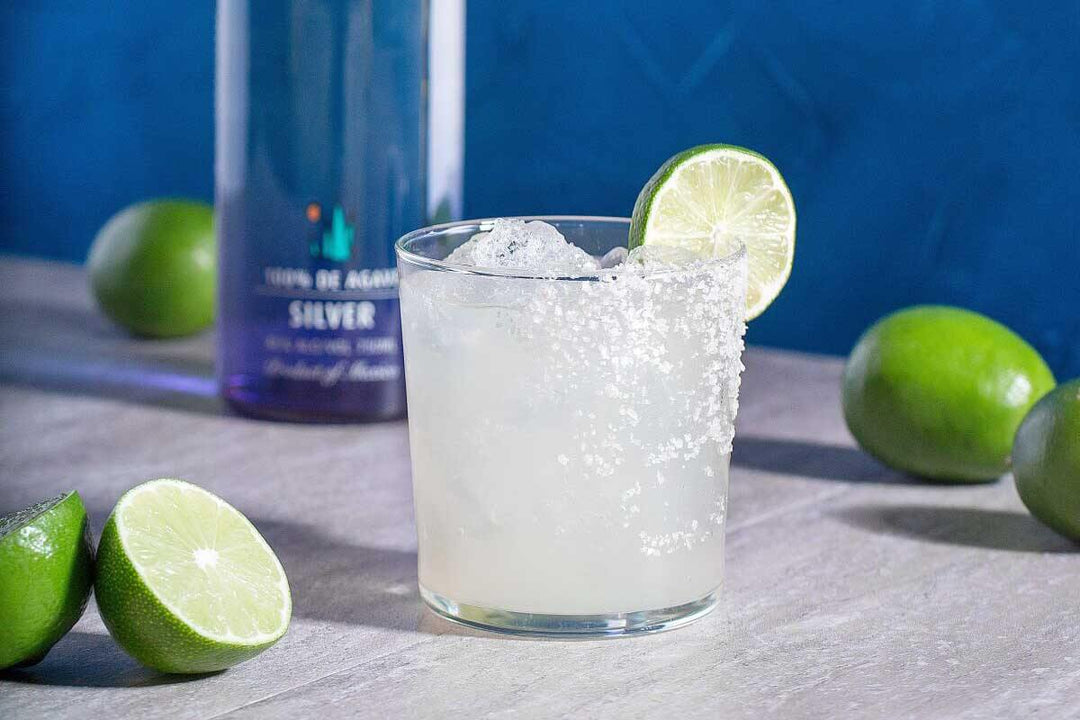 A glass of margarita with a salted rim and a lime slice on the rim is on a table. Limes and a bottle are in the background against a blue backdrop.