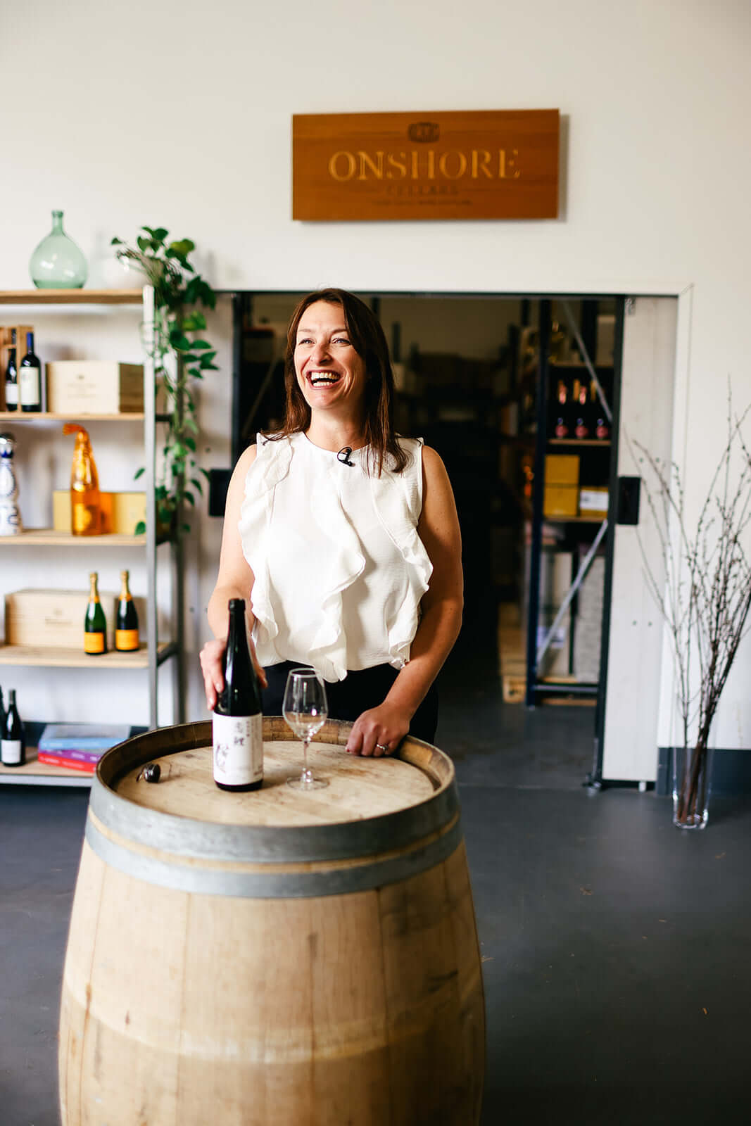 A woman laughing stands behind a wooden wine barrel with a wine bottle and glass. Shes inside a wine shop with shelves of bottles and a sign that reads ONSORE. The atmosphere is casual and inviting.