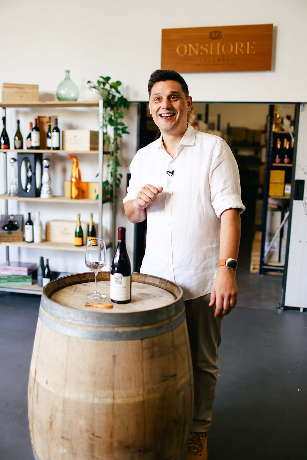 A person in a white shirt stands smiling behind a wine barrel with a bottle and glass of red wine. The background features a shelf with wine bottles and a sign reading Onshore Cellar.