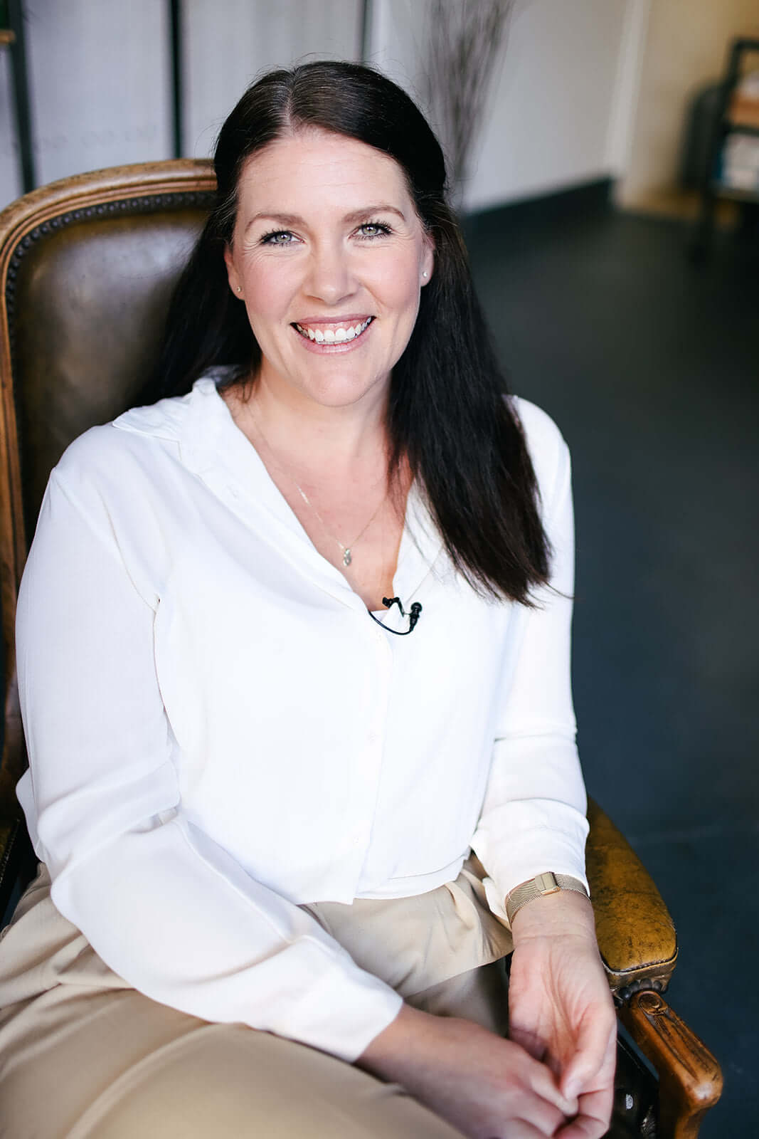 Smiling woman with long dark hair sits in a brown leather chair, wearing a white blouse and beige pants. She is indoors, in a room with a dark floor and light walls.