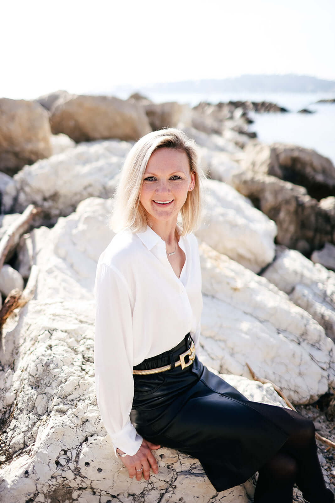 A person with blonde hair sits on large white rocks by the sea, wearing a white long-sleeve shirt and a dark skirt. The background features more rocks and calm water under a bright sky.