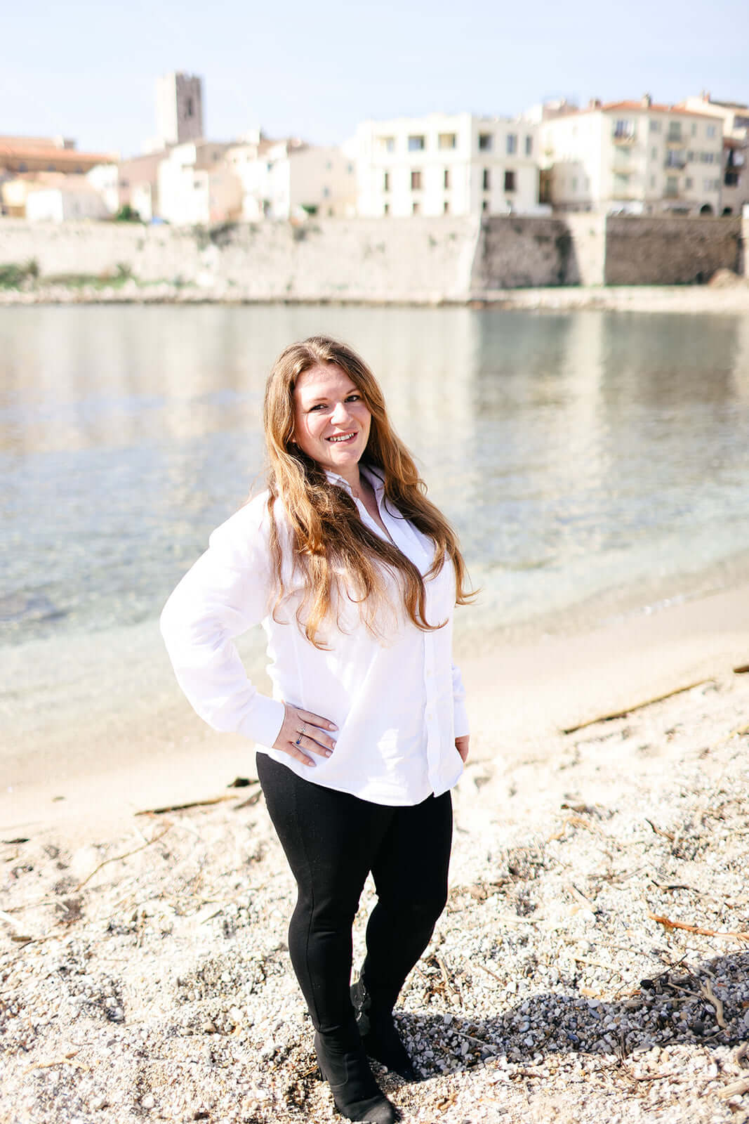 A woman with long hair stands on a pebble beach, wearing a white top and black pants. She is smiling with her hand on her hip. The background shows calm water and buildings along the shoreline under a clear sky.