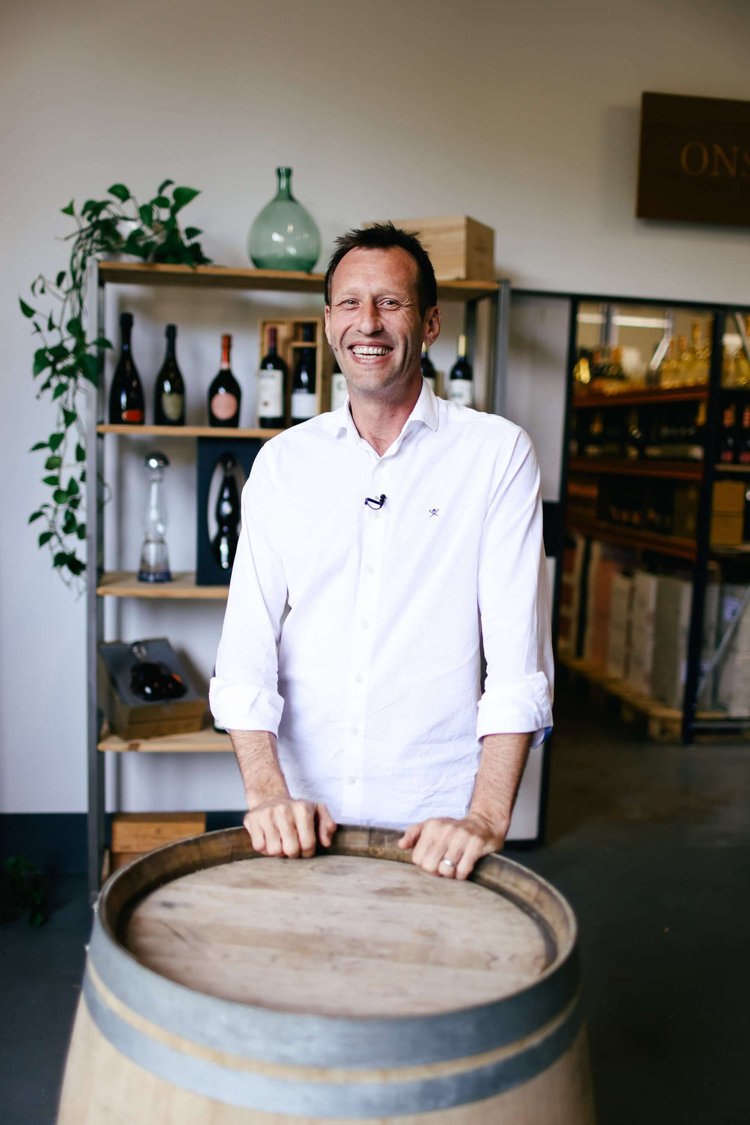 A man in a white shirt stands smiling behind a wooden barrel in a wine shop. Shelves with various wine bottles and a plant are in the background.