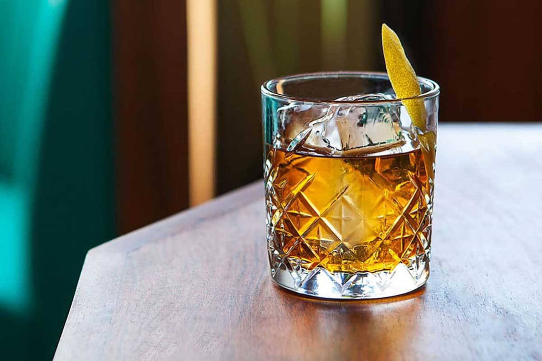 A crystal glass filled with an amber-colored cocktail on a wooden table, garnished with a twist of lemon peel. The background is softly blurred, giving a warm and inviting atmosphere.