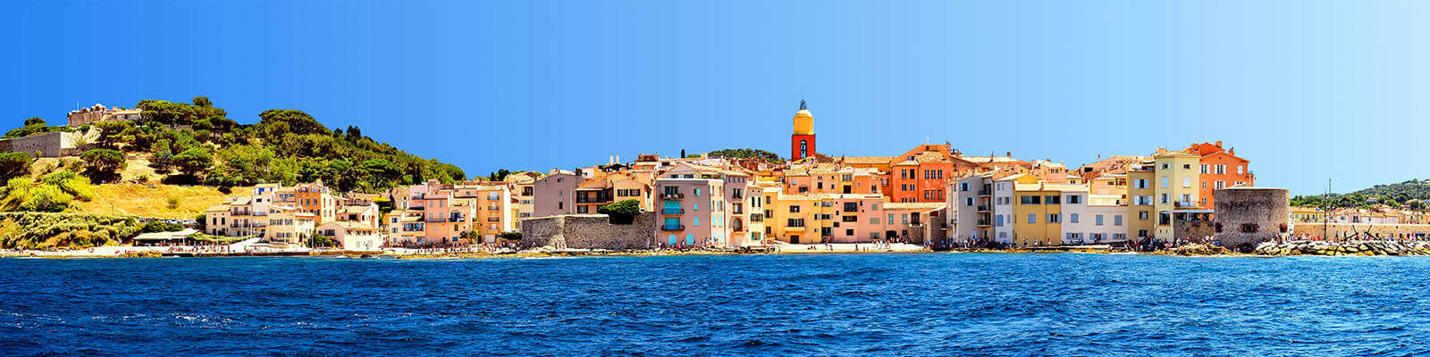 Panoramic view of a coastal town with colorful buildings along the shoreline. A prominent bell tower rises in the center. The sea is a vivid blue, and greenery is visible on a hillside to the left under a clear sky.