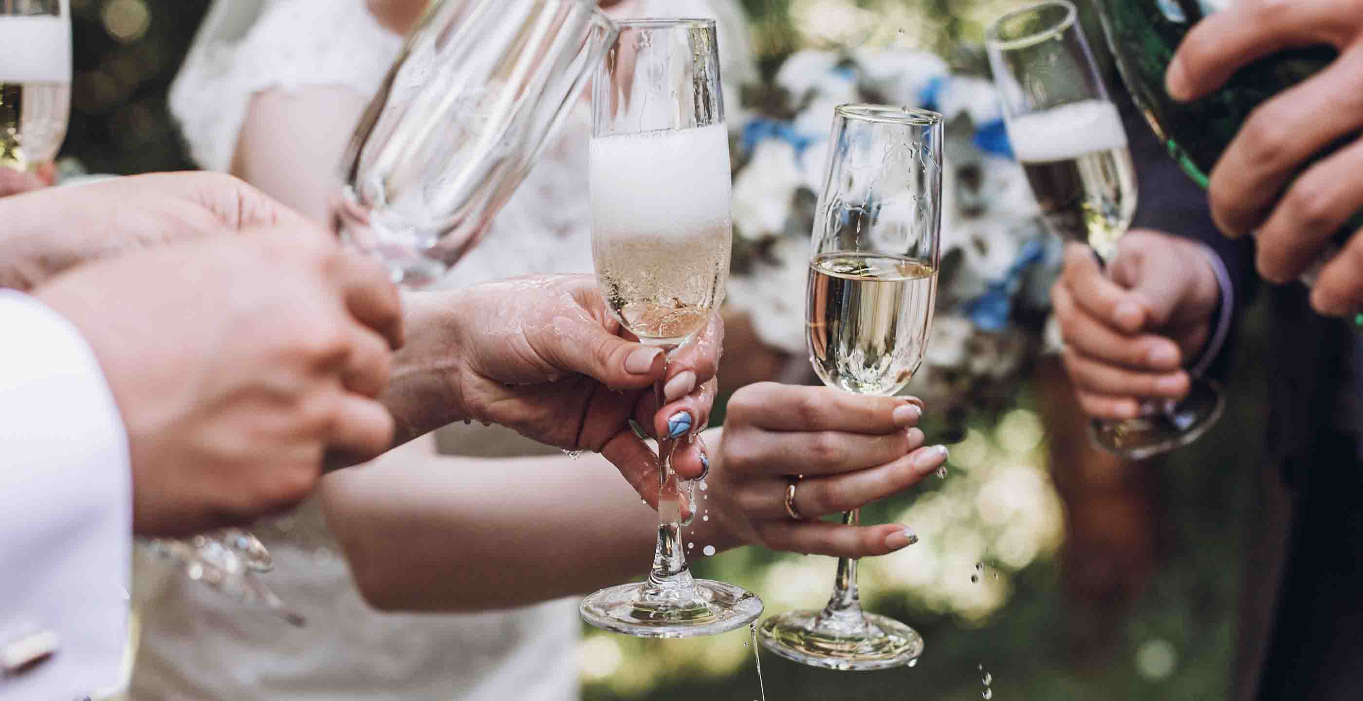 A group of people clinking champagne glasses together in a celebratory toast. The background shows a blurred outdoor setting with greenery.