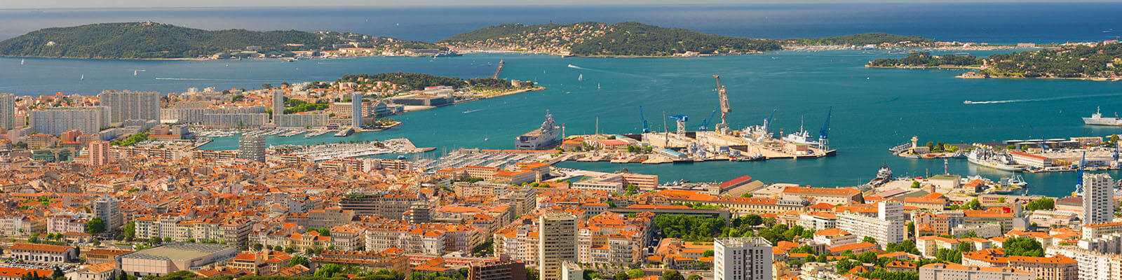 Aerial view of a coastal city with orange-roofed buildings by the turquoise sea, featuring ships docked at a busy port. Islands with lush greenery are visible in the background under a clear blue sky.