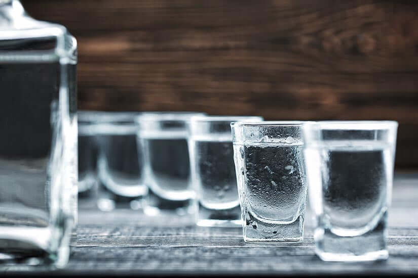 A row of shot glasses filled with clear liquid, possibly vodka, sits on a wooden surface. A blurred bottle is visible on the left. The background is a rustic wooden texture.