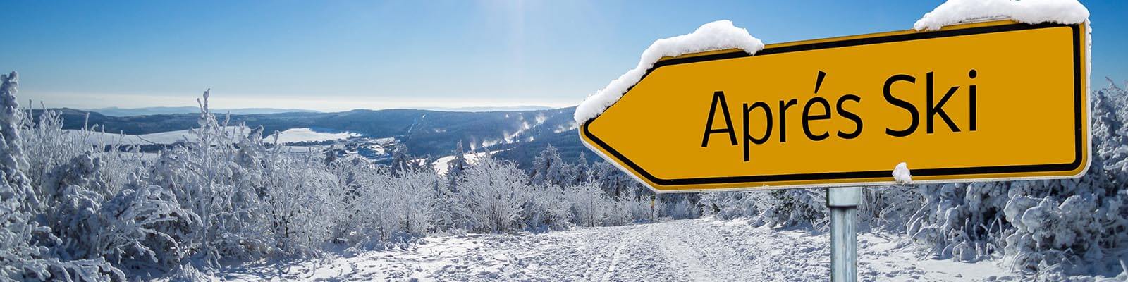 A bright yellow road sign labeled Après Ski is covered with snow and points toward a snow-covered landscape. The background features snow-laden trees under a clear blue sky, with distant hills visible on the horizon.