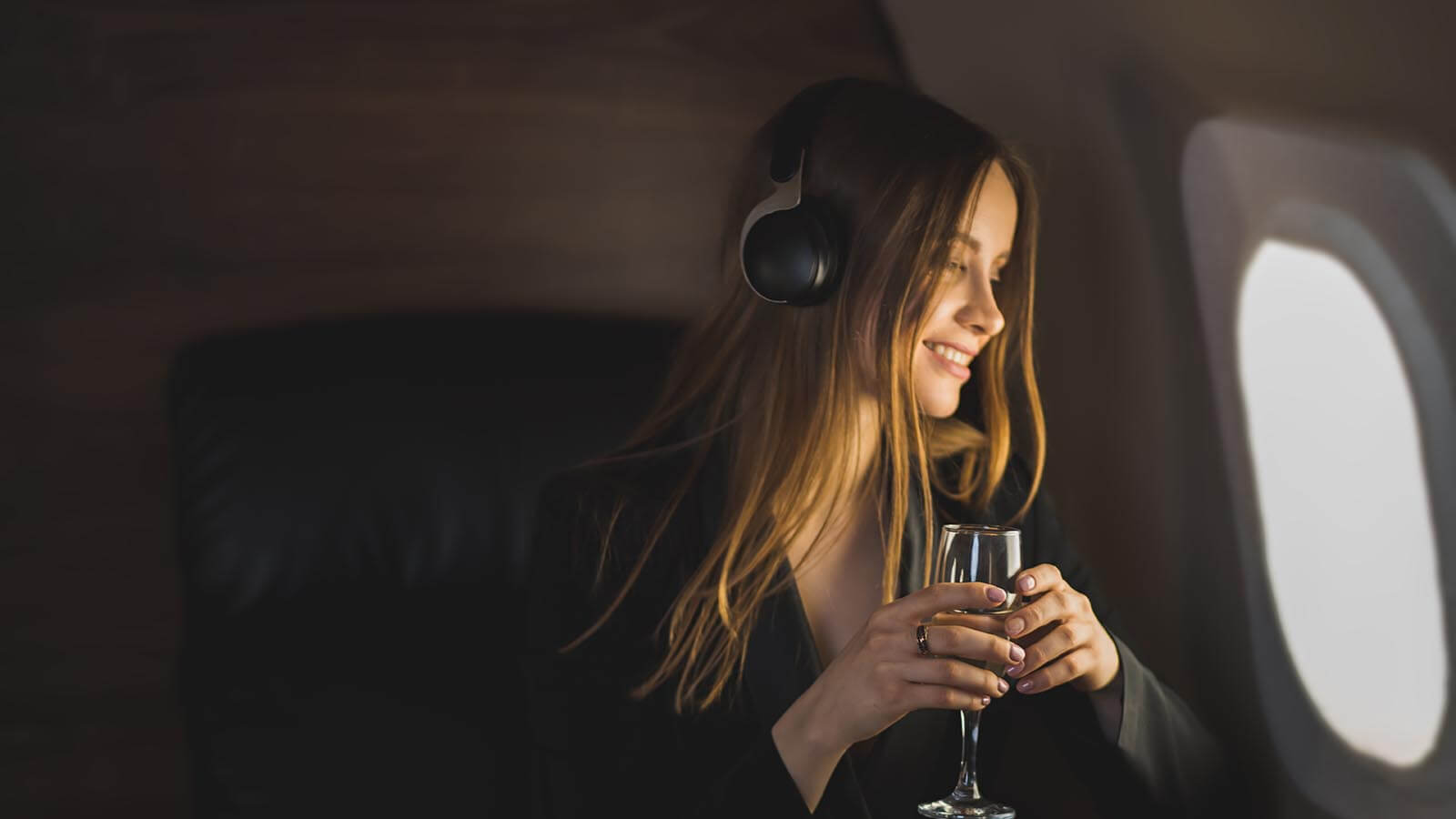 A woman wearing headphones is sitting in an airplane seat, smiling while looking out the window. She is holding a glass, and the background shows the interior of the plane.
