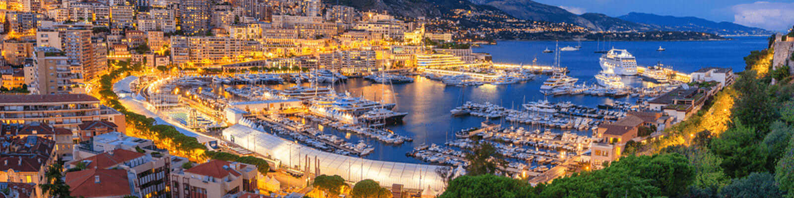 A panoramic view of a marina at dusk, with numerous yachts docked in the harbor. The surrounding cityscape is illuminated, creating a warm, golden glow against the blue water and sky. Hills and mountains provide a scenic backdrop.
