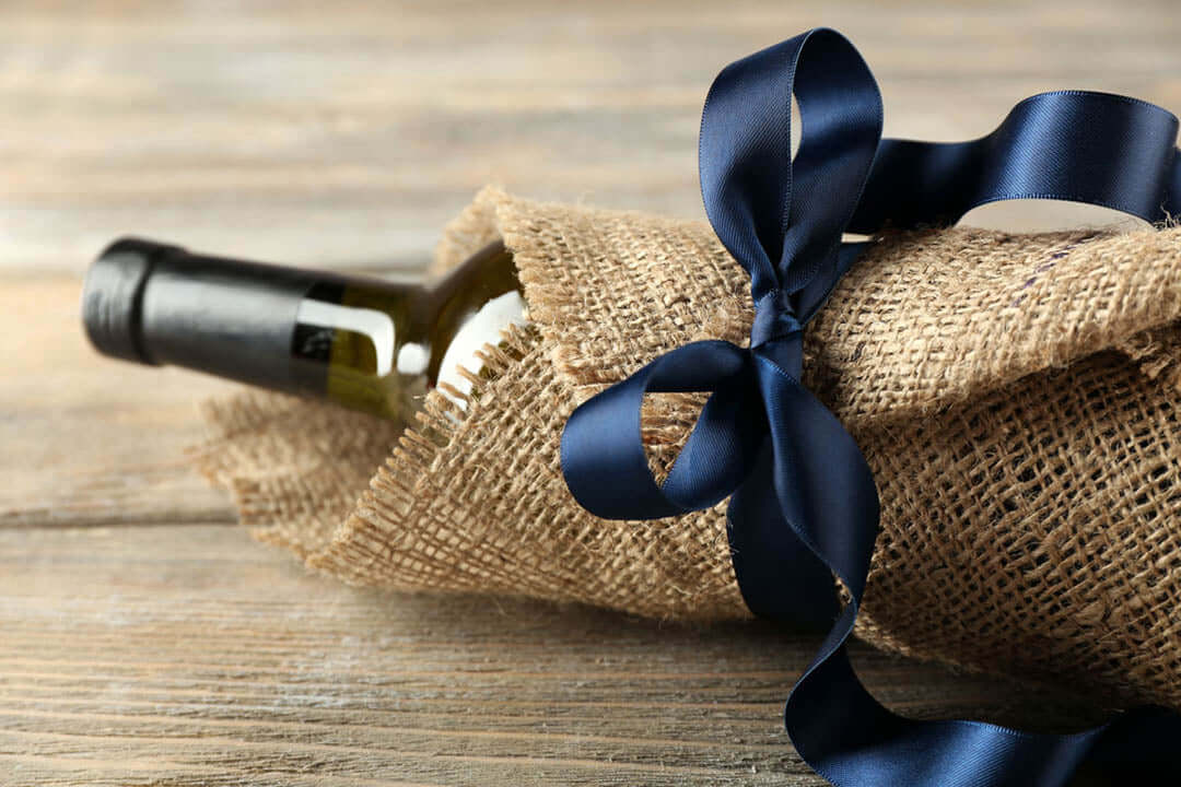A wine bottle wrapped in burlap fabric is tied with a navy blue satin ribbon. The bottle is laying on a wooden surface.