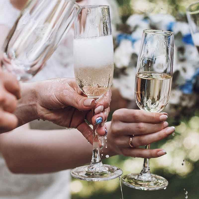 Close-up of hands toasting with champagne flutes outdoors. The glasses are filled with bubbly champagne, and some liquid is spilling. The background is blurred, with hints of greenery and white flowers.